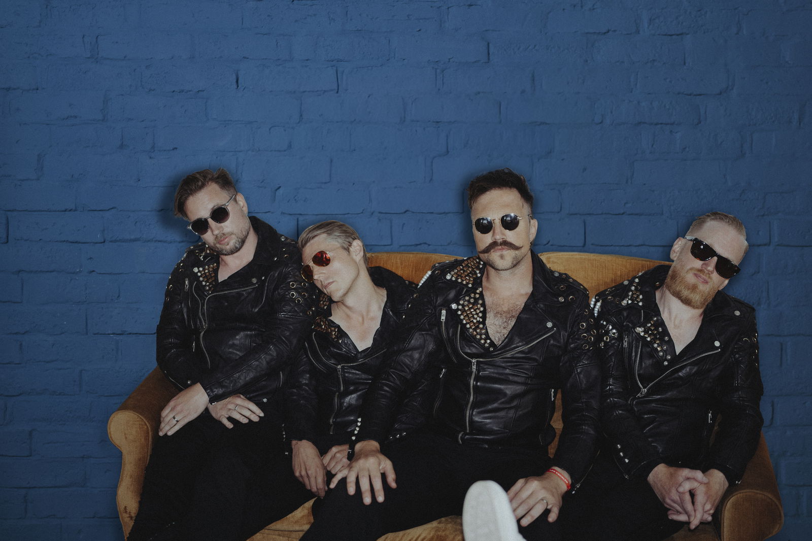 Royal Republic sitzen auf deinem gelb-braunen Samt-Sofa vor einer knallblauen Ziegelsteinmauer